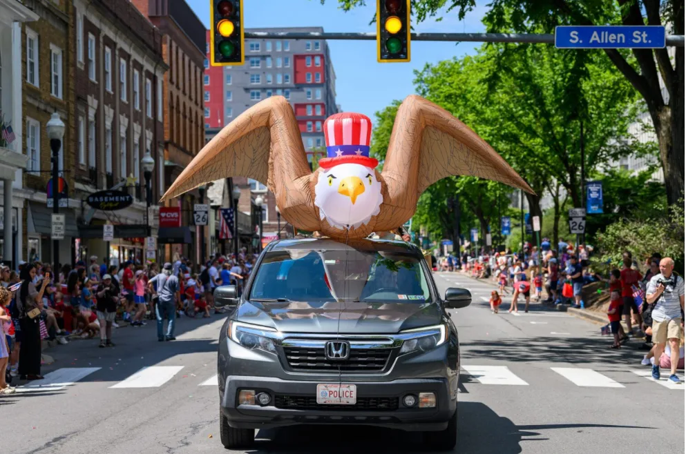 An inflatable bald eagle wearing an Uncle Sam hat rides atop a truck down South Allen Street, with crowds lining the sidewalks, during the 2026 Independence Day Parade in State College